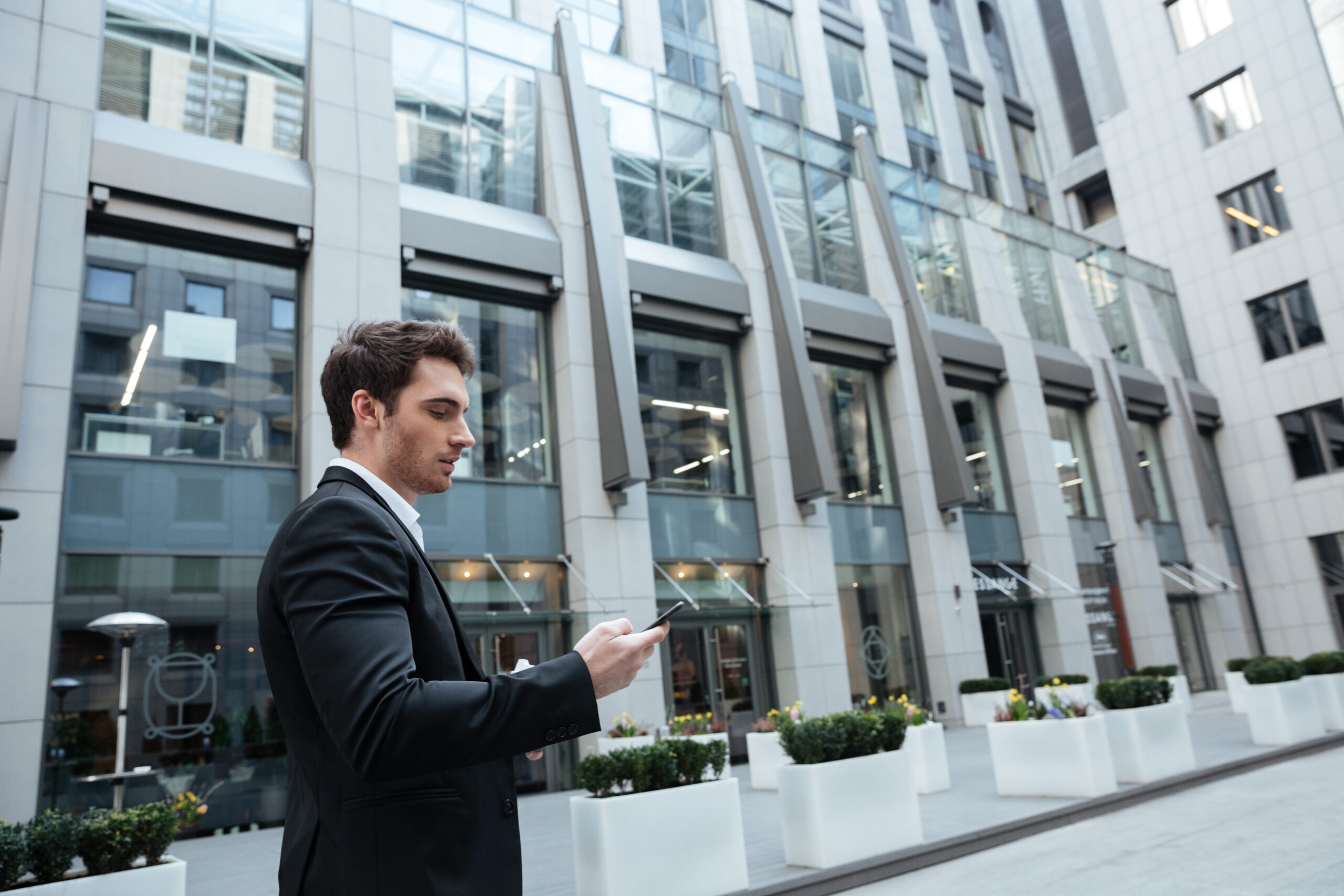 A man in a black suit stands outdoors in a modern city business district, looking at his smartphone. Tall glass-and-steel office buildings rise behind him, and white planter boxes with greenery line the walkway